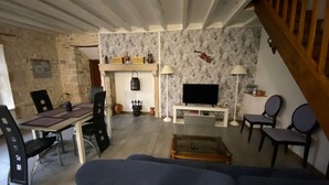 Ein Wohnzimmer mit einer Couch, Fernseher und Kamin. Es gibt einen Essbereich mit Stühlen und einem Tisch. Der Raum hat Steinwände und eine Holzbalkendecke.
