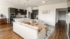 A modern living room with a large sectional sofa, wooden flooring, and a kitchen in the background. - House in North Fort Worth (Haslet)