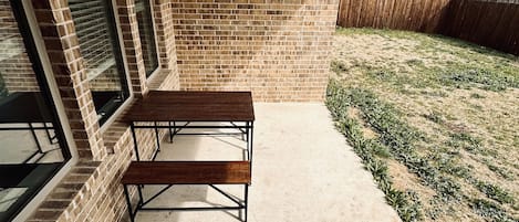 A covered patio area with a brick wall, a table and bench, and a view of a grassy area with a wooden fence.