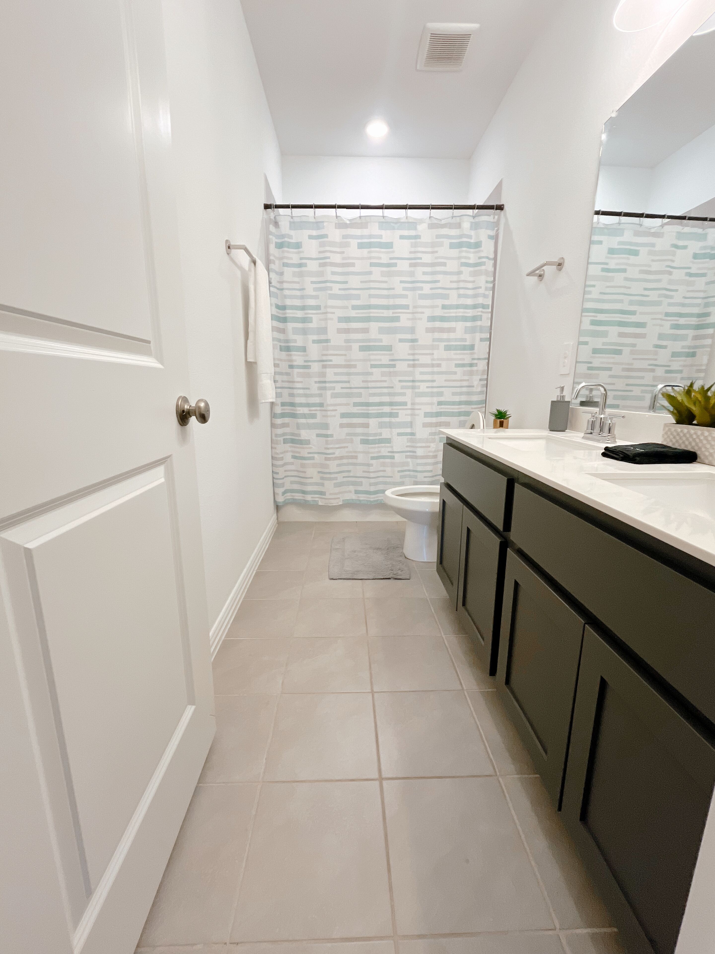 A modern bathroom with a white door, a shower curtain with a geometric pattern, a toilet, a sink with a mirror, and a small plant on the counter.