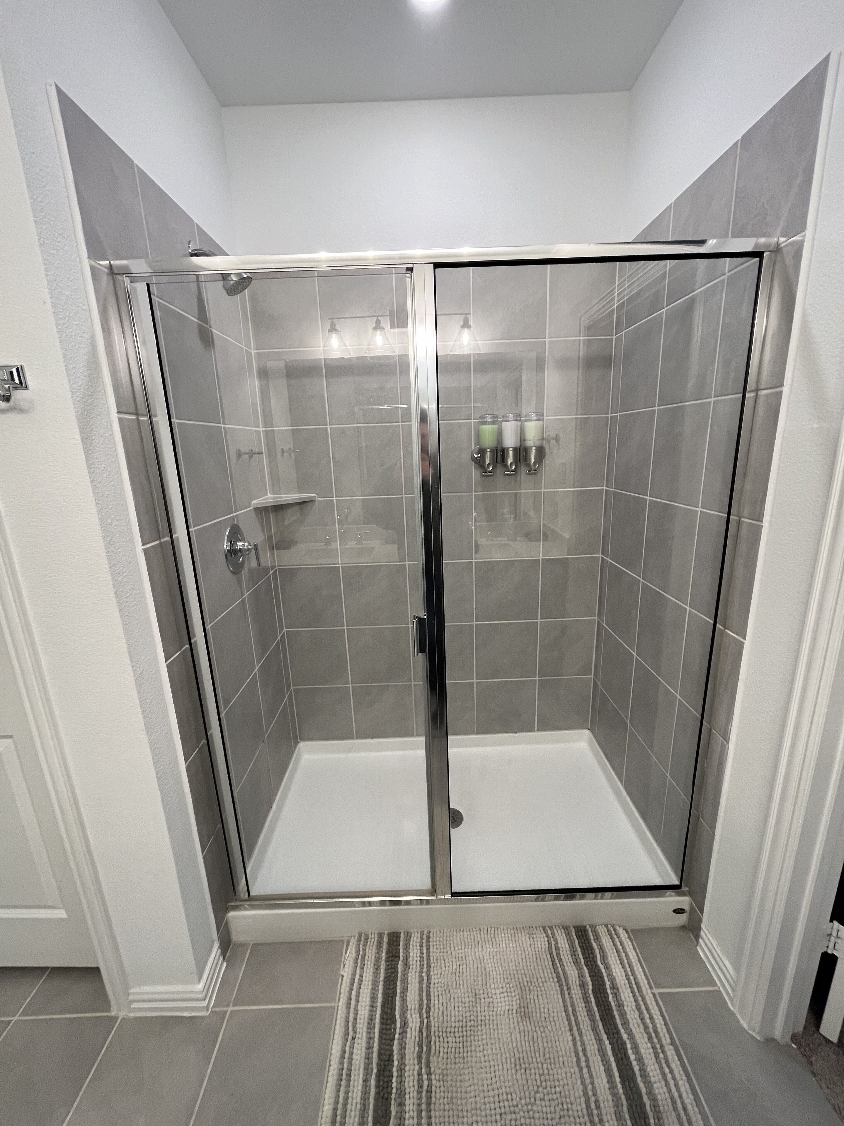 A modern shower enclosure with a glass door, a tiled floor, and a small rug.