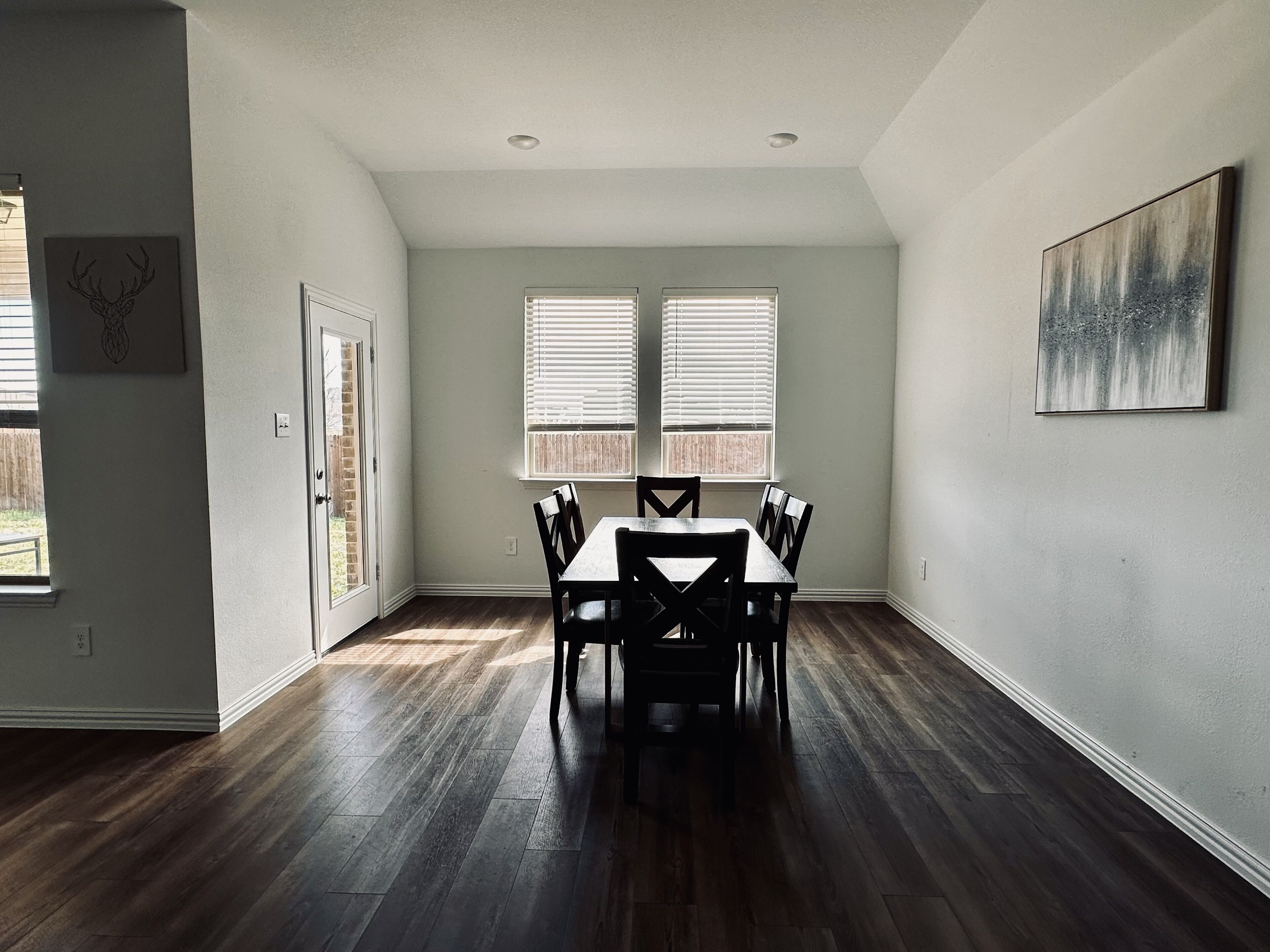 A dining area with a table and chairs, two windows with blinds, and a painting on the wall.
