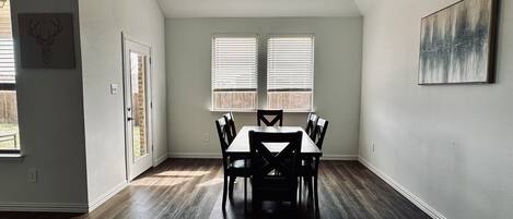 A dining area with a table and chairs, two windows with blinds, and a painting on the wall.
