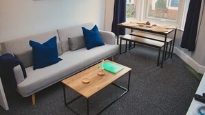 Una sala moderna con un sofá gris, dos cojines azules, una mesa de centro de madera y una pequeña mesa con un mantel tejido y un cuenco.