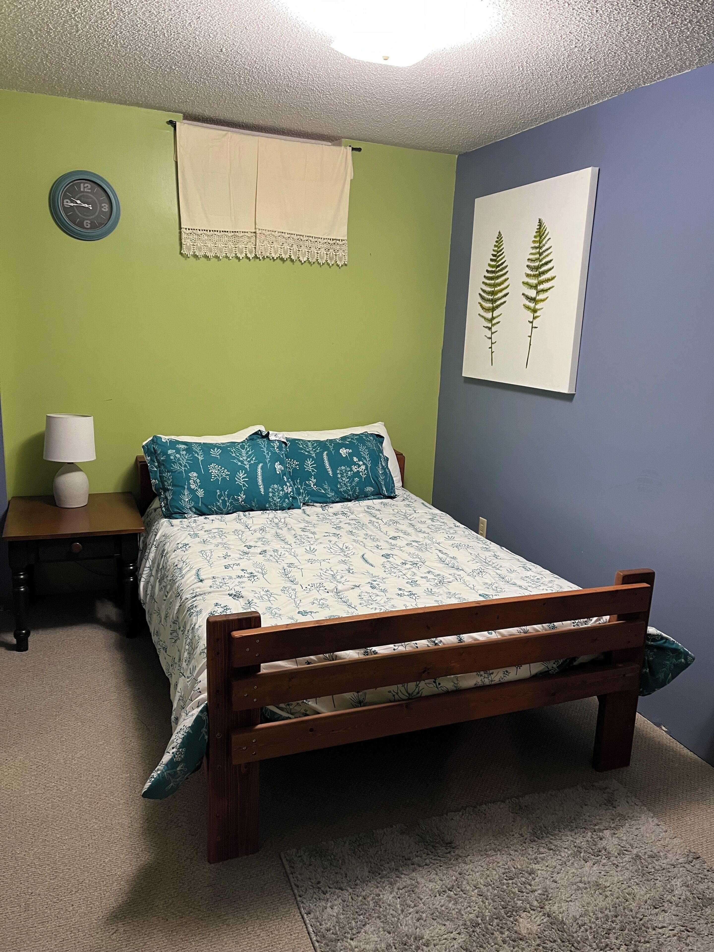 A bedroom with a wooden bed, a nightstand, a lamp, a clock, and a framed plant.