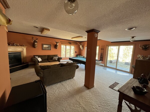 A spacious living room with a fireplace, a pool table, and a large window offering a view of the outdoors.