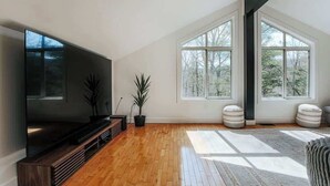 Ein geräumiges Wohnzimmer mit großem Flachbildfernseher, Holzboden und großen Fenstern mit Blick auf Bäume.