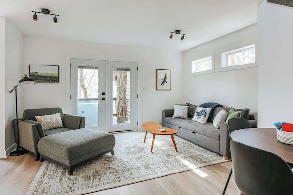 Un salon moderne avec un canapé gris, un pouf assorti, une table basse en bois et un lampadaire. Il y a deux fenêtres avec des rideaux, une porte donnant sur l’extérieur et un tableau encadré au mur.