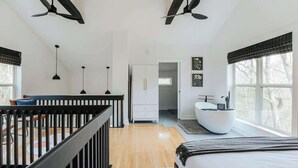 A modern bedroom with a wooden floor, a bed, a white bathtub, and a desk with a chair. There are two ceiling fans and a window with a view of trees. - The Nest · Hot Tub, Forest Views & Soaking Tub (Berkeley Springs)
