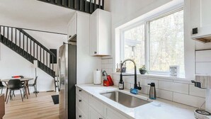 A modern kitchen with white cabinetry, a stainless steel refrigerator, a sink, and a window with a view of trees. - The Nest · Hot Tub, Forest Views & Soaking Tub (Berkeley Springs)