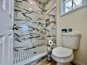 A bathroom with a toilet, a shower with a marble wall, and a window.