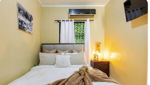 Une chambre à coucher comprenant un lit, une table de chevet avec une lampe, une fenêtre avec des rideaux et un appareil de climatisation.