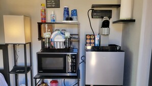 A compact kitchenette with a microwave, toaster, and refrigerator. There are shelves with dishes, a coffee maker, and a small table with a cup of coffee.