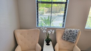 Two beige armchairs with a small table between them, a potted plant, and a window with blinds.