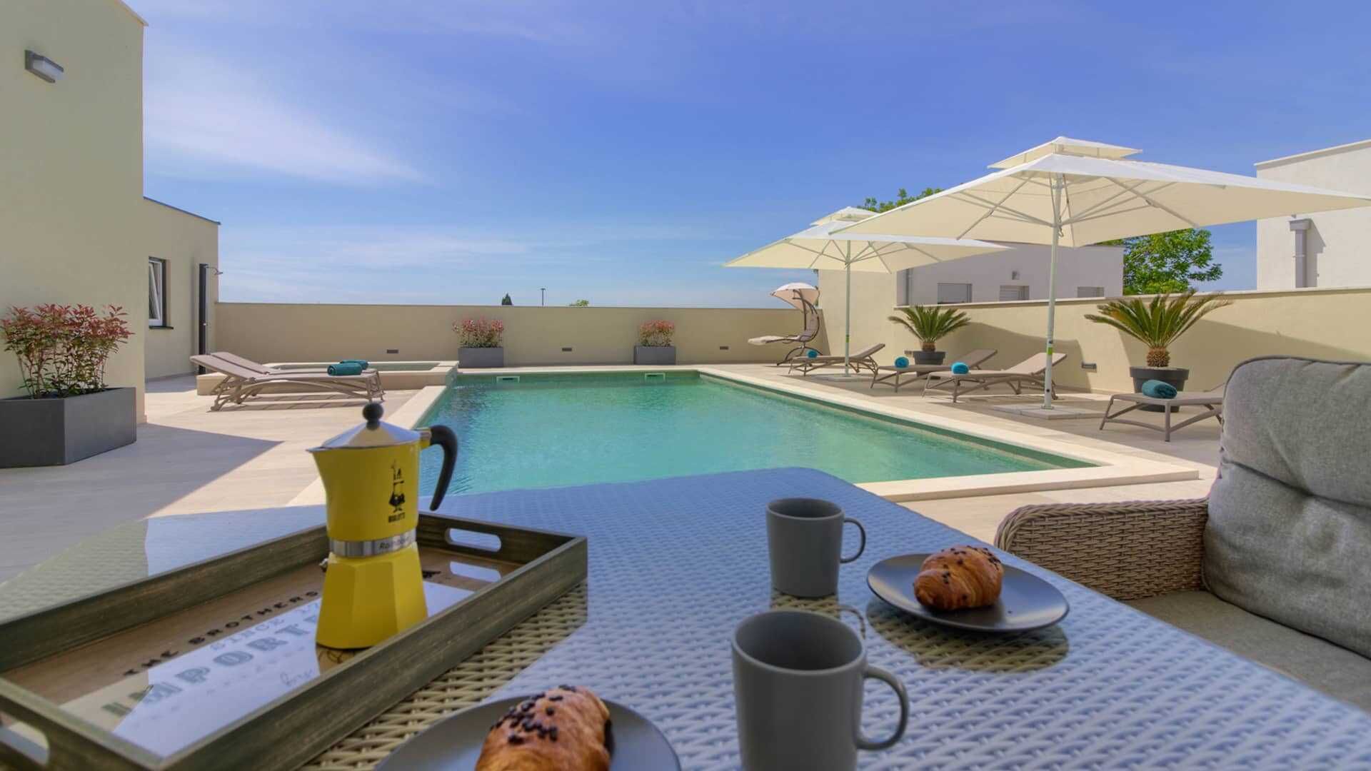 A poolside area with a table set for breakfast, including a croissant and a mug, and a coffee pot.