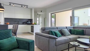 Un espacio habitable moderno con un sofá gris, cojines turquesa, una pequeña mesa redonda y una zona de cocina con armarios blancos y encimera negra.