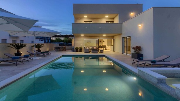 Un área de piscina moderna al aire libre con sillones de descanso, zona de comedor y un edificio de dos pisos con amplias ventanas.