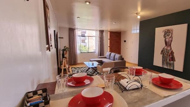 Un comedor con una mesa lista para cuatro personas, un cuadro en la pared y una vista de una sala de estar al fondo.