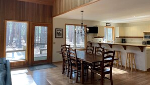 A spacious dining area with a wooden table and chairs, a kitchen with white cabinets, and large windows offering a view of the forest. - Woodzy Retreat Getaway (Black Butte Ranch)