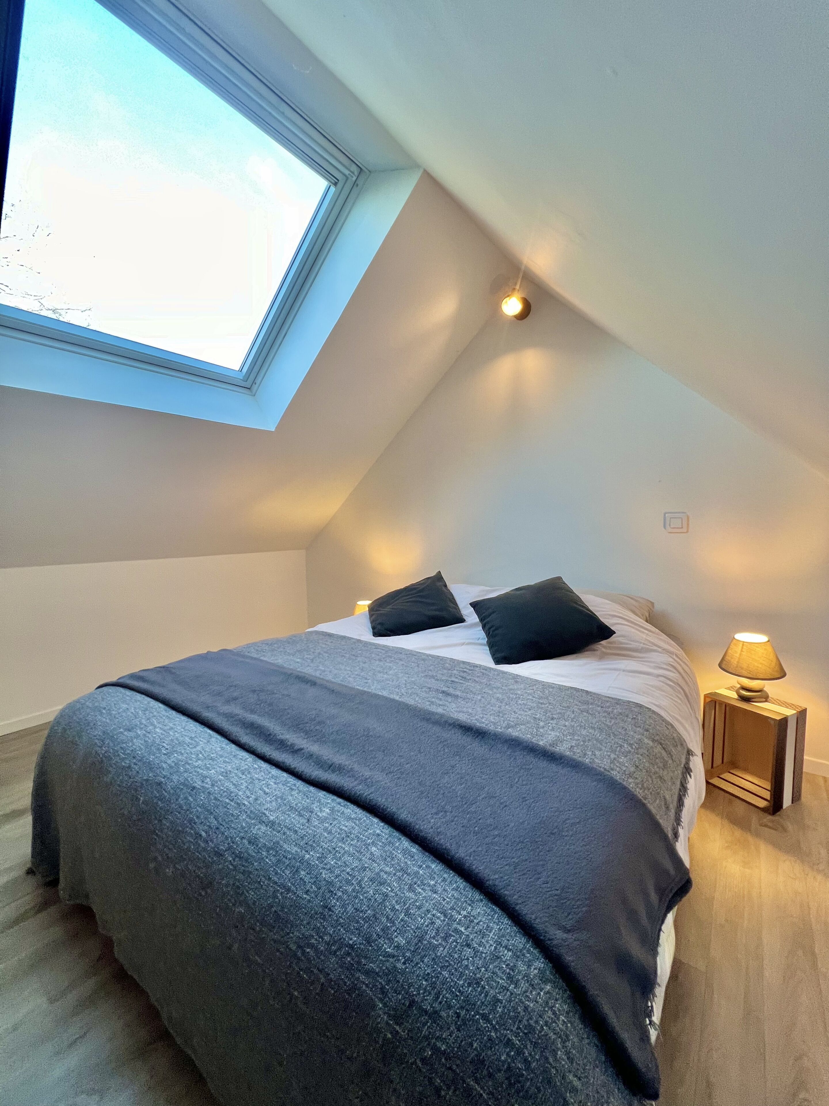 Une chambre avec une lucarne, un lit recouvert d’une couverture grise, des oreillers noirs et une lampe de chevet.