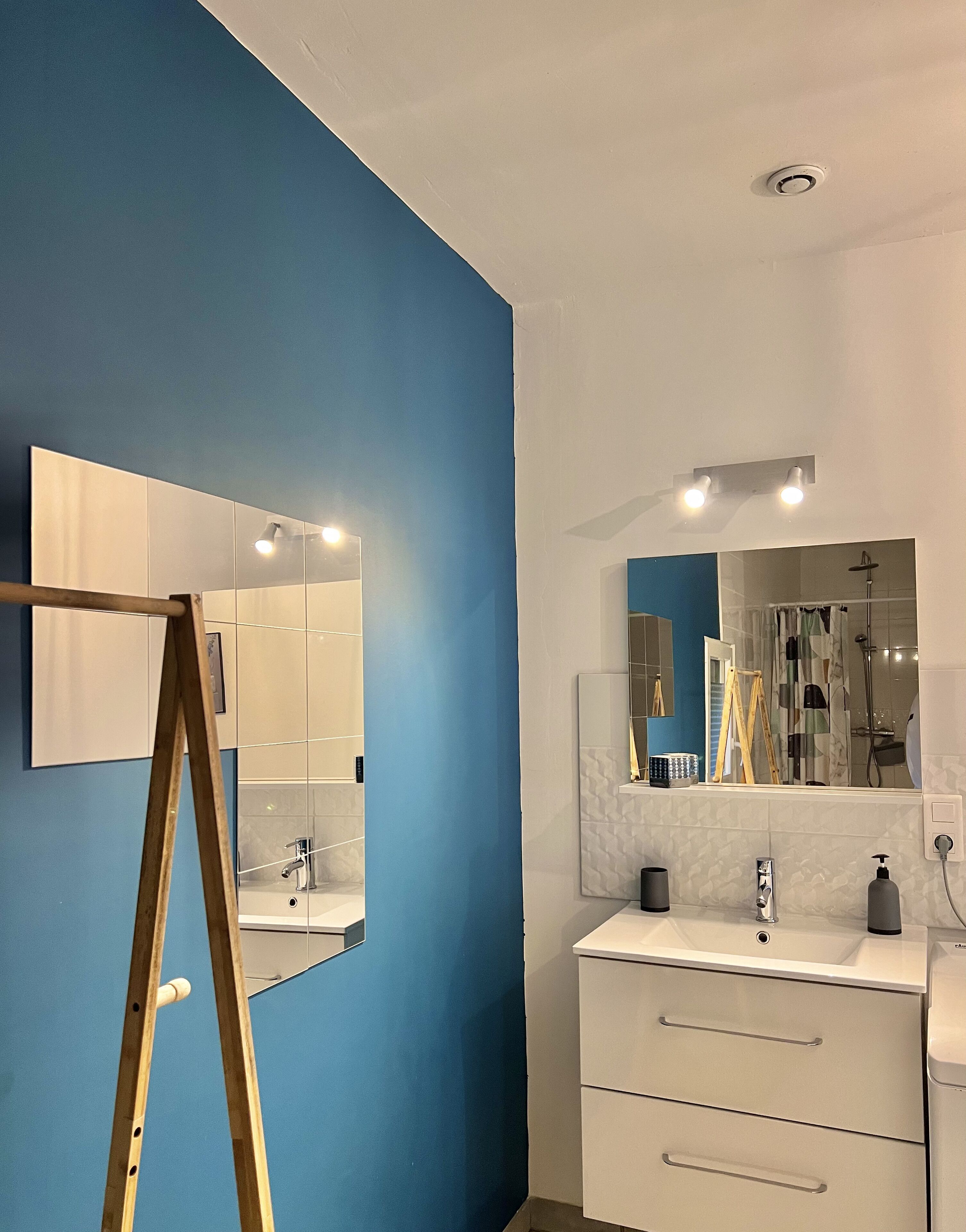 Une salle de bain avec un mur bleu, un chevalet en bois, un grand miroir, un lavabo blanc et un espace douche.
