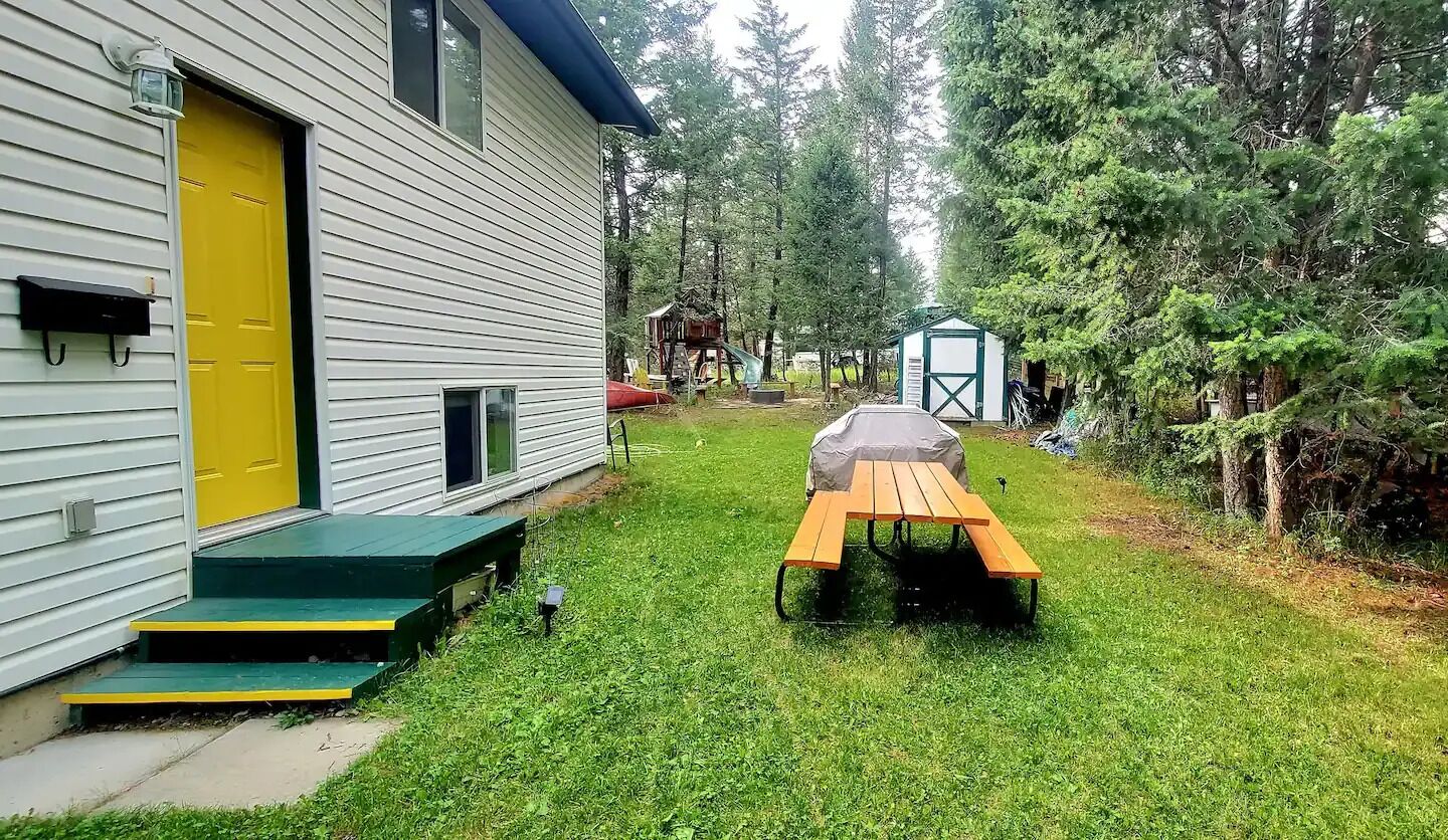 A house with a yellow door, a picnic table, and a small shed in a grassy yard surrounded by trees.