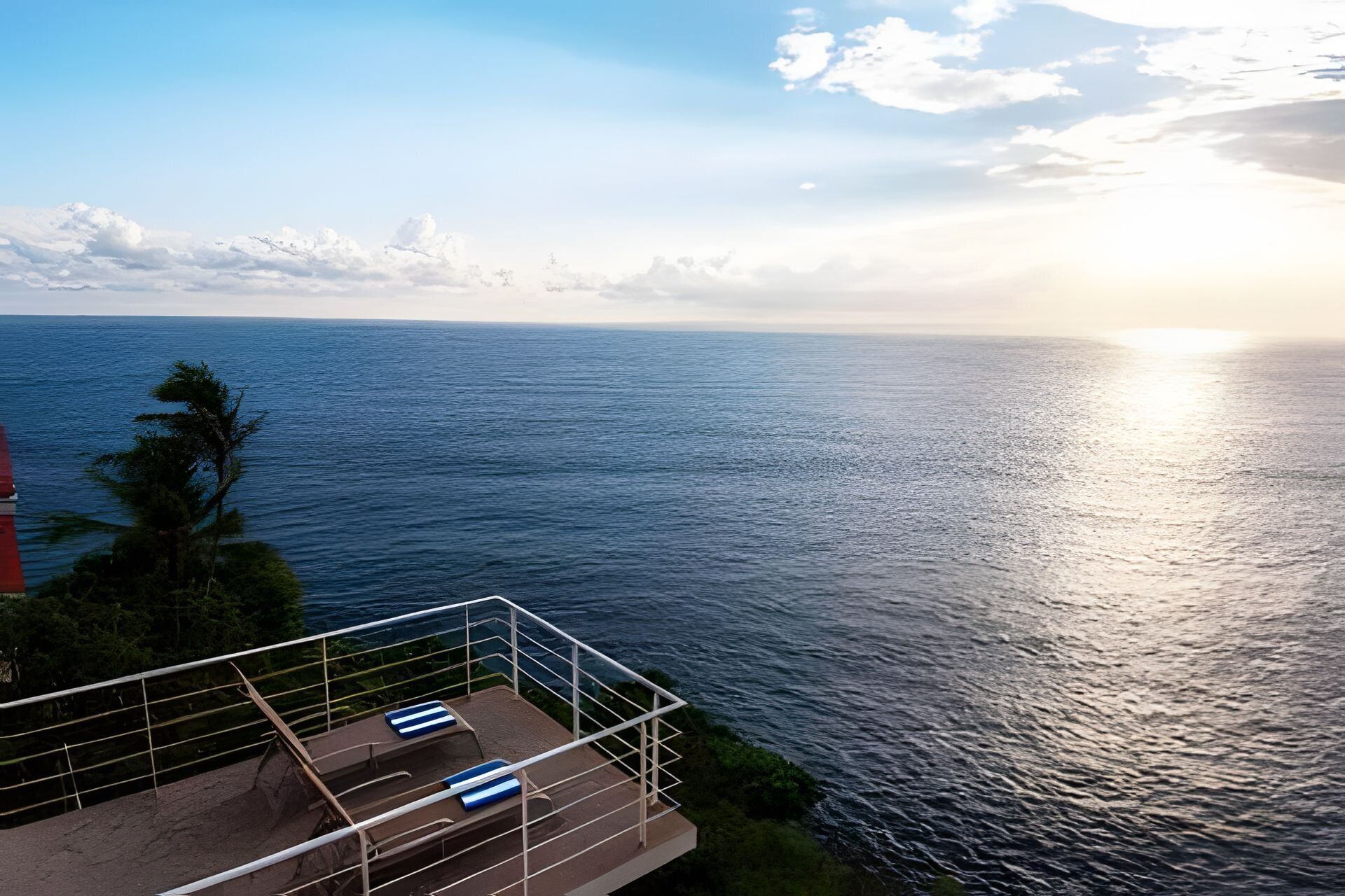 Un balcón con dos sillones reclinables a rayas azules, con vista a un mar tranquilo durante el atardecer.