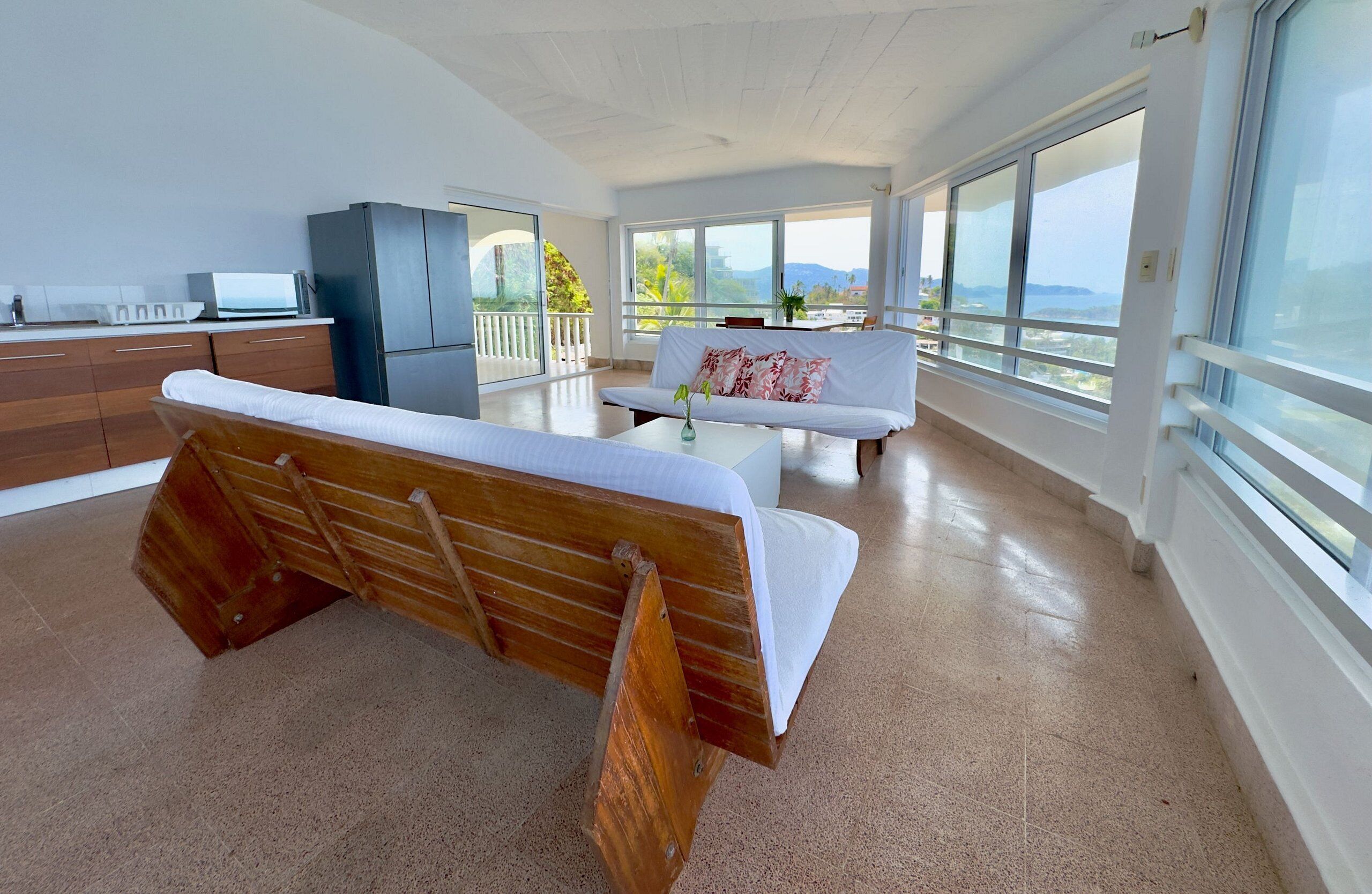 Een moderne woonkamer met een houten bank, een witte bank, een eettafel met een vaas, en grote ramen met uitzicht.