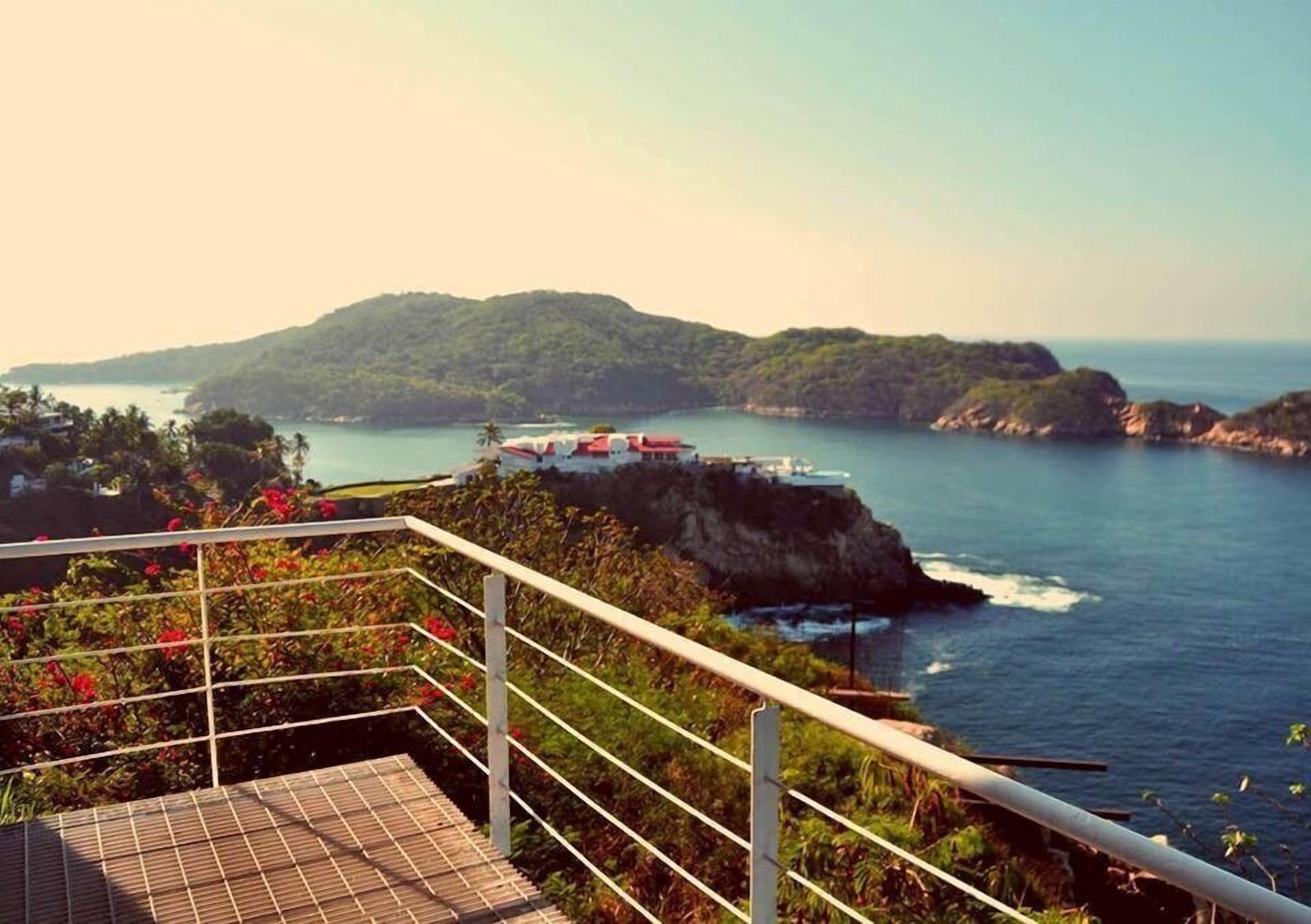 Vista costera con una escalera que conduce a una terraza con vistas al mar, un pequeño edificio de techo rojo y abundante vegetación.