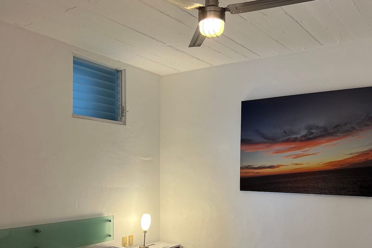 Un dormitorio con cama, ventilador de techo, mesita de noche con lámpara, ventana con persianas y un cuadro de un atardecer.