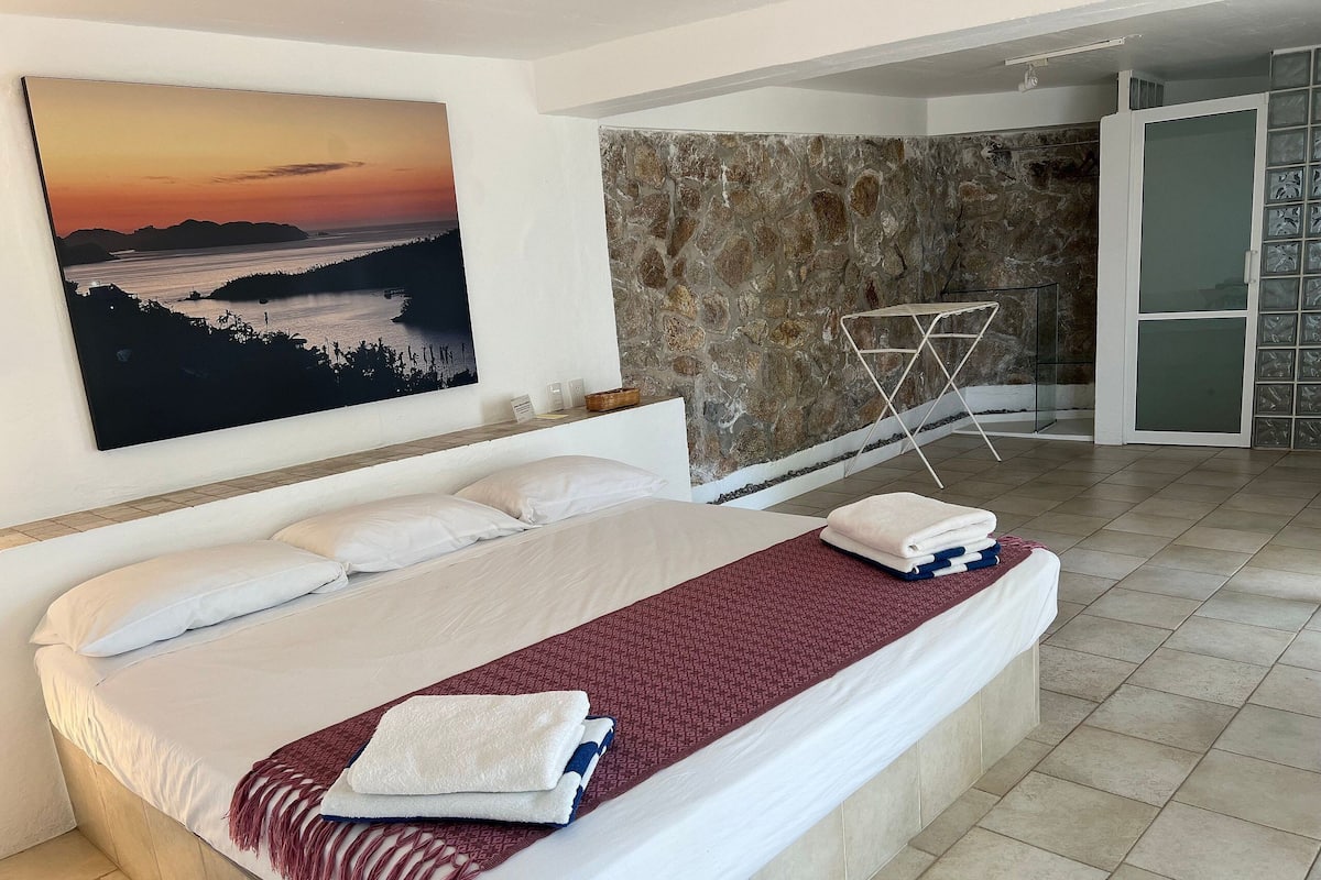 Una cama bien hecha con sábanas blancas y una manta roja, una pared de piedra y un cuadro de un atardecer.