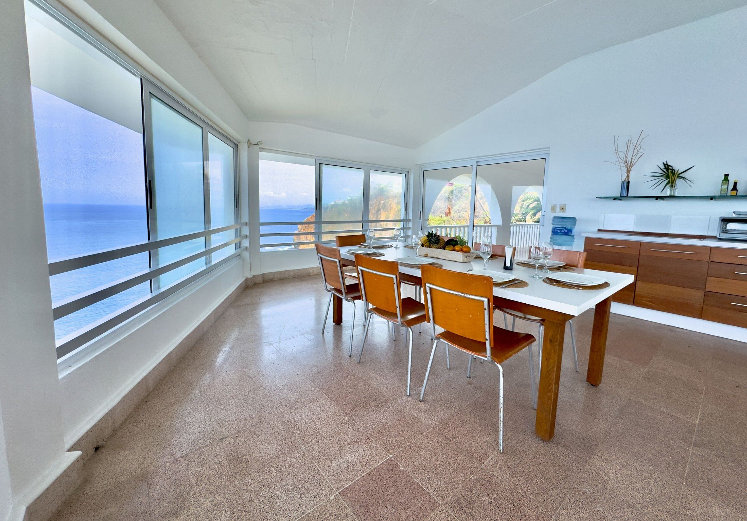 Una cocina moderna con ventanas amplias, un comedor con sillas y vista al mar.