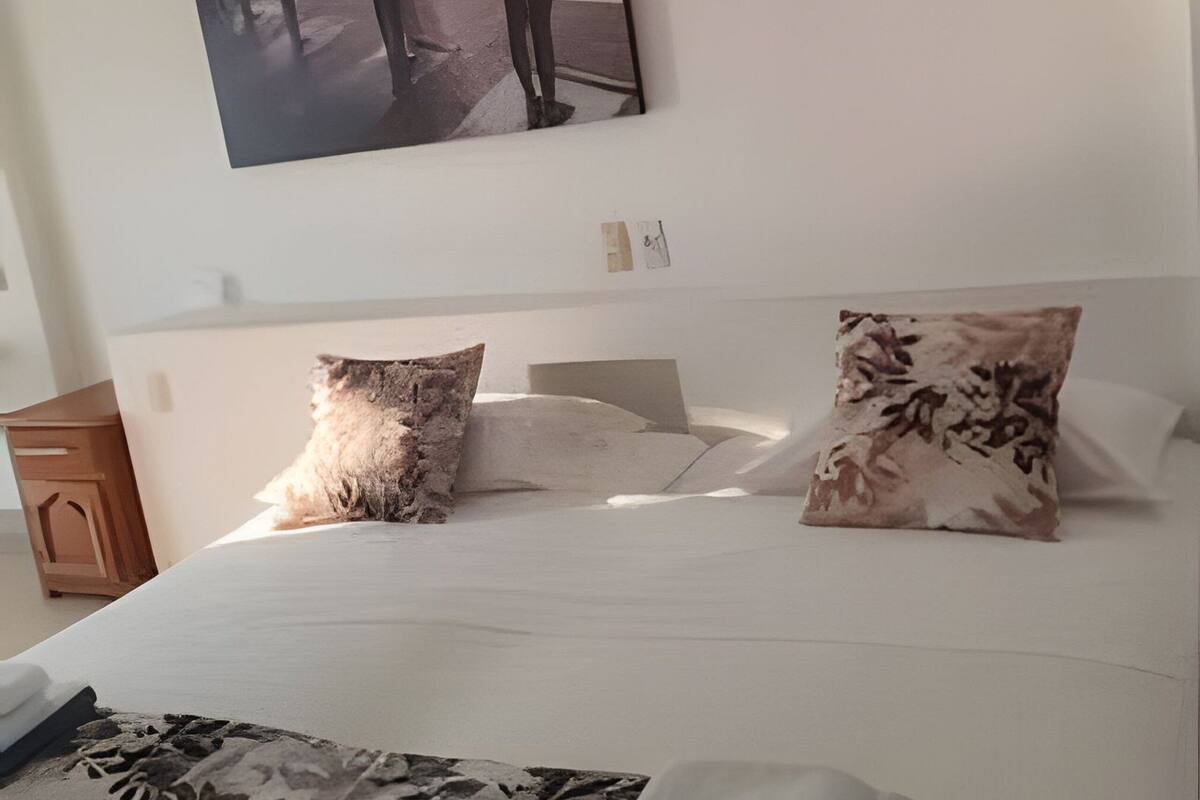 Un dormitorio con cama, almohadas, una mesita de noche y una obra de arte colgada en la pared.