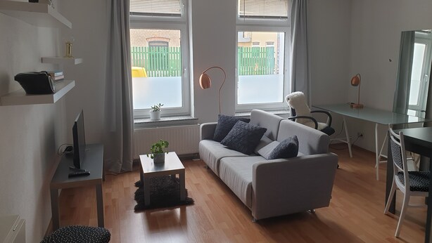 A modern living room with a grey sofa, a small table, a desk with a chair, and a large window with curtains. - 2 Zimmer-wohnung am Landesmuseum für Vorgeschichte (Halle)