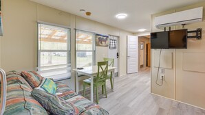 A compact living space with a sofa, a dining table with chairs, a television mounted on the wall, and an air conditioning unit. - Horse Farm Cottage Near WEC + Rainbow Springs (Morriston)