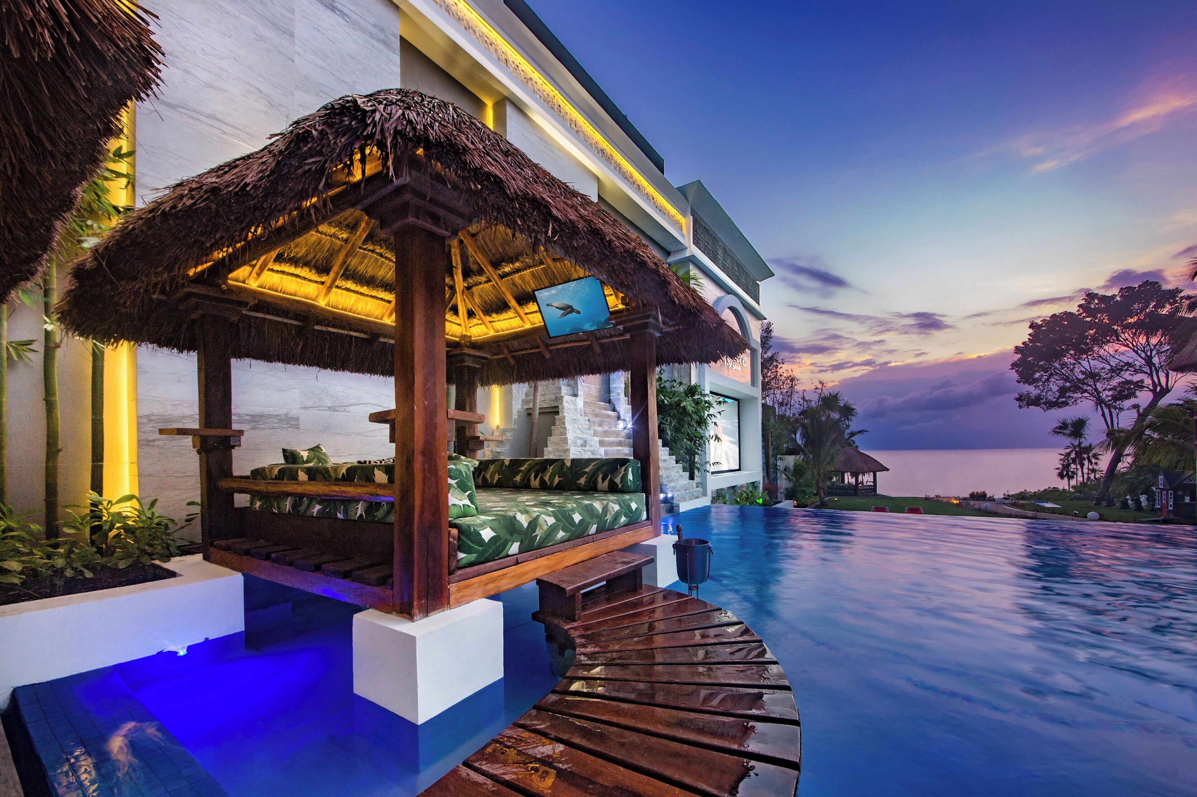 A luxurious resort with a private infinity pool, a thatched-roof lounge area, and a view of the ocean at dusk.