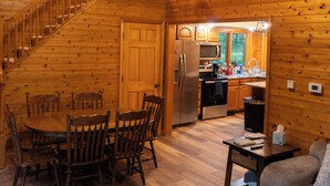 A wooden cabin interior with a dining area, kitchen, and a staircase leading to an upper level.
