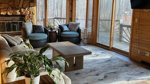 A cozy cabin interior with a stone fireplace, wooden ceiling, large windows, and a stone wall.