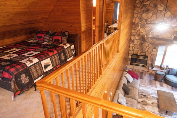 Interior acolhedor de uma cabana com escada de madeira, lareira de pedra, sofá e cama com colcha estampada.