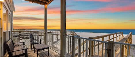 A wooden deck with outdoor seating, overlooking a beach with a wooden railing.