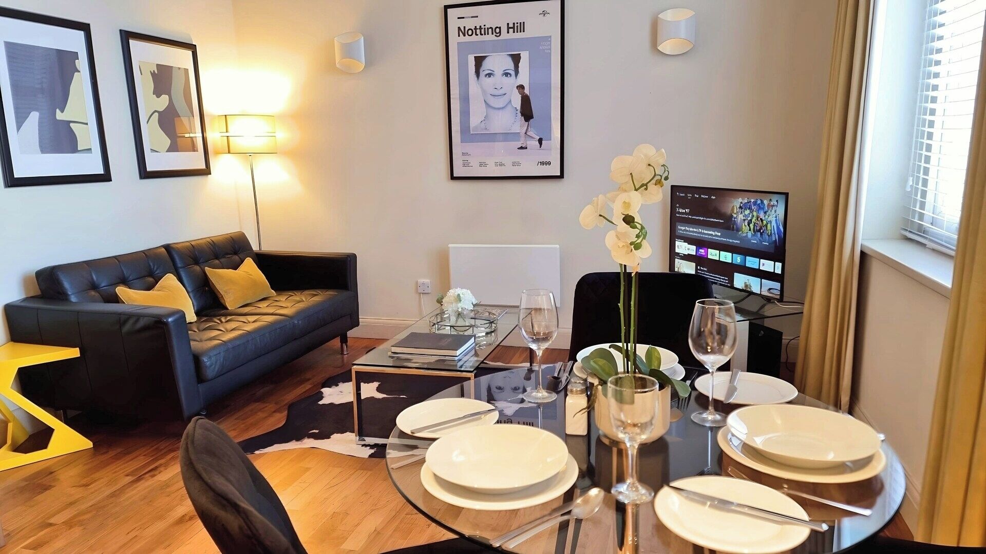 A modern living room with a black leather sofa, a glass coffee table, and a dining table set for four. There are framed pictures on the wall, a poster, and a TV displaying a music video.