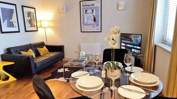 A modern living room with a black leather sofa, a glass coffee table, and a dining table set for four. There are framed pictures on the wall, a poster, and a TV displaying a music video.