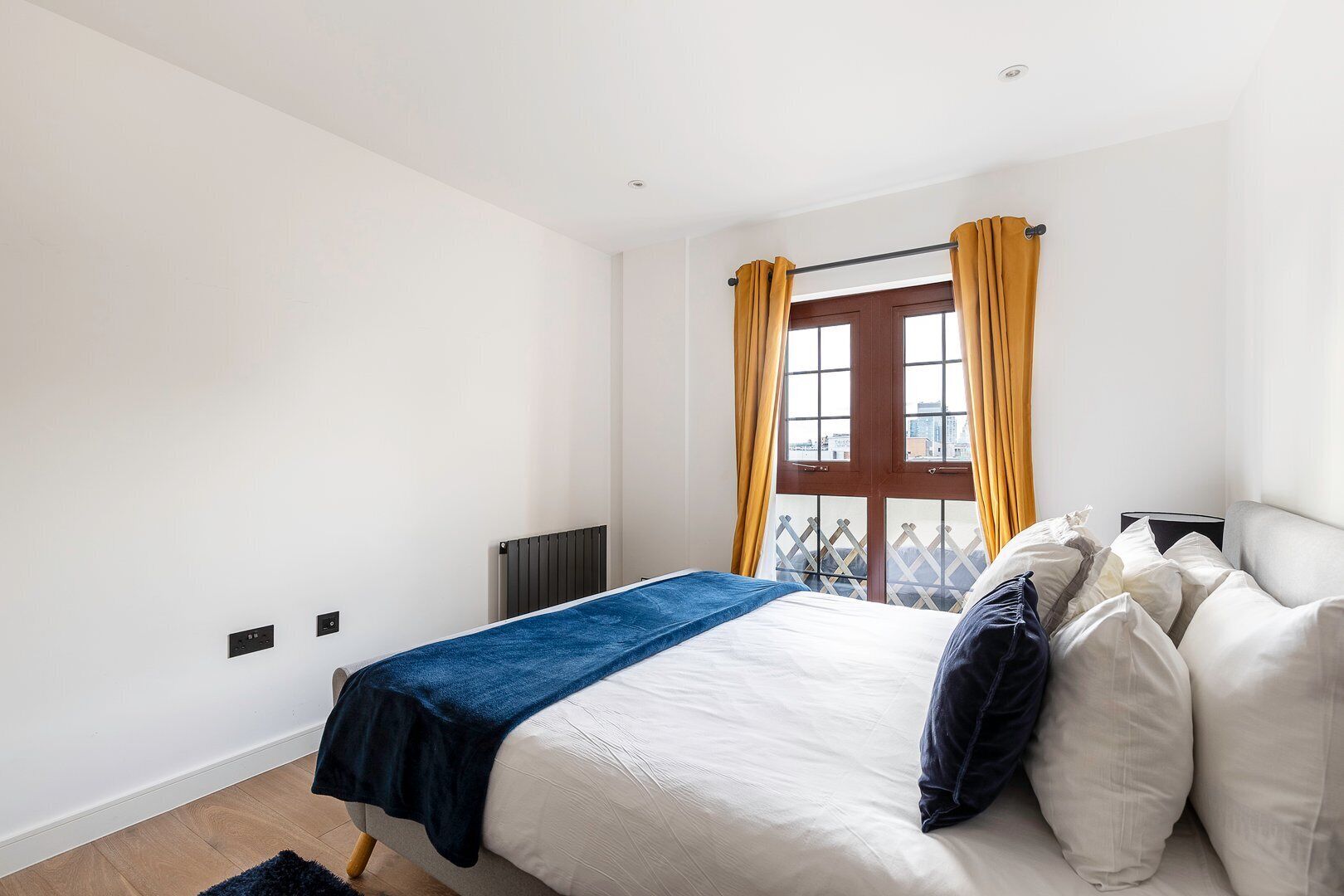 A bedroom with a bed, a radiator, a window with curtains, and a view of the city.