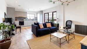 A modern living room with a dark blue sofa, a glass coffee table, and a kitchen area in the background. - City View Spacious 2 Bedroom Penthouse (London)