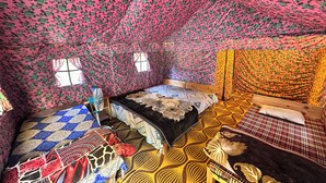 A room with a sloped ceiling, floral patterned walls, a bed with a patterned bedspread, a small table with a fan, and a window. - The Gazzaar Riverside camp (Nainital)