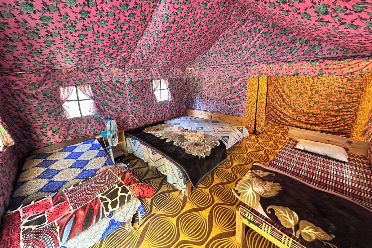 A room with a sloped ceiling, floral patterned walls, a bed with a patterned bedspread, a small table with a fan, and a window.