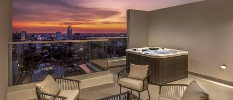 A balcony with a hot tub, wicker chairs, and a city view at sunset.