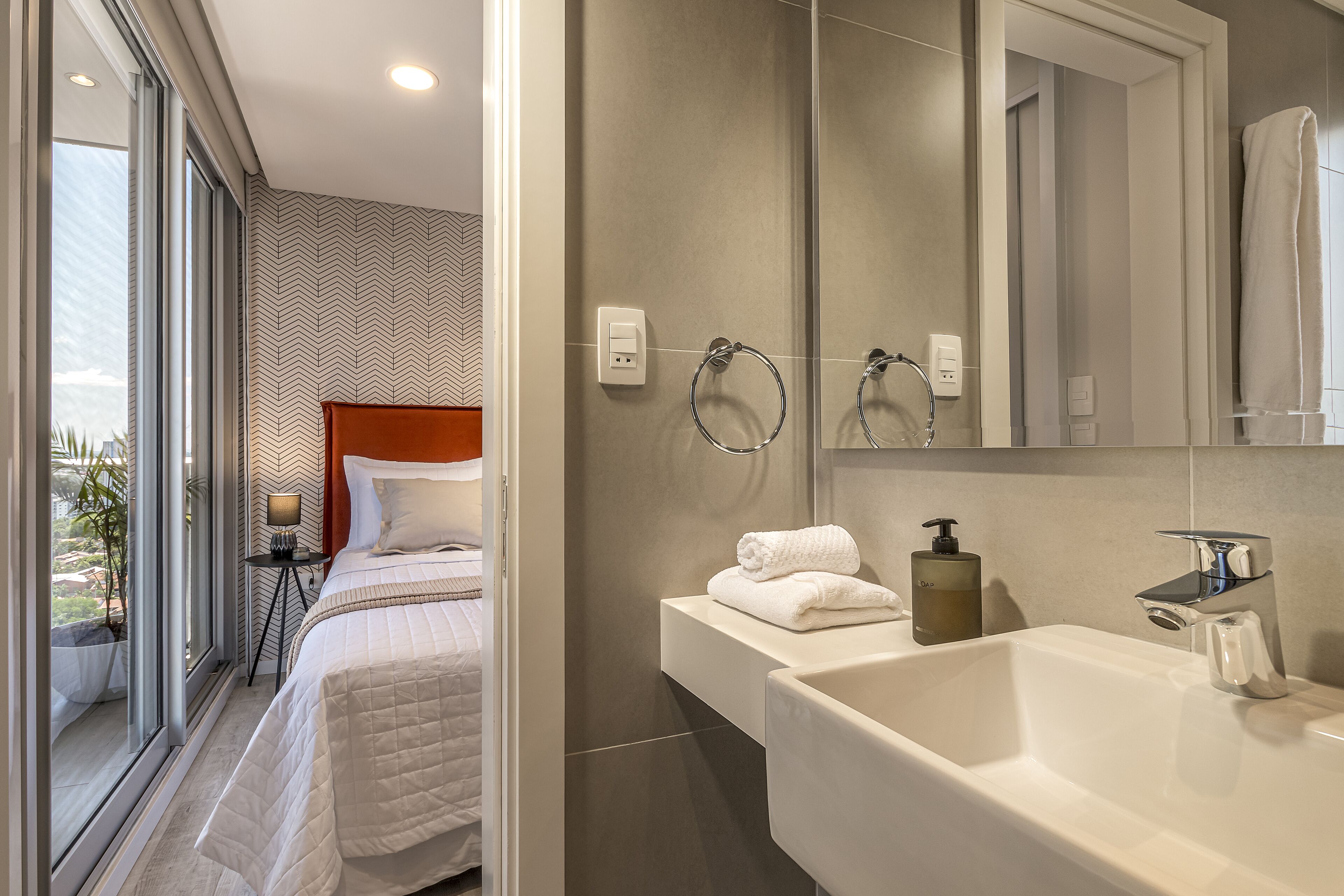 Una habitación de hotel moderna con una cama grande, un baño con lavamanos y espejo, y vistas al exterior.