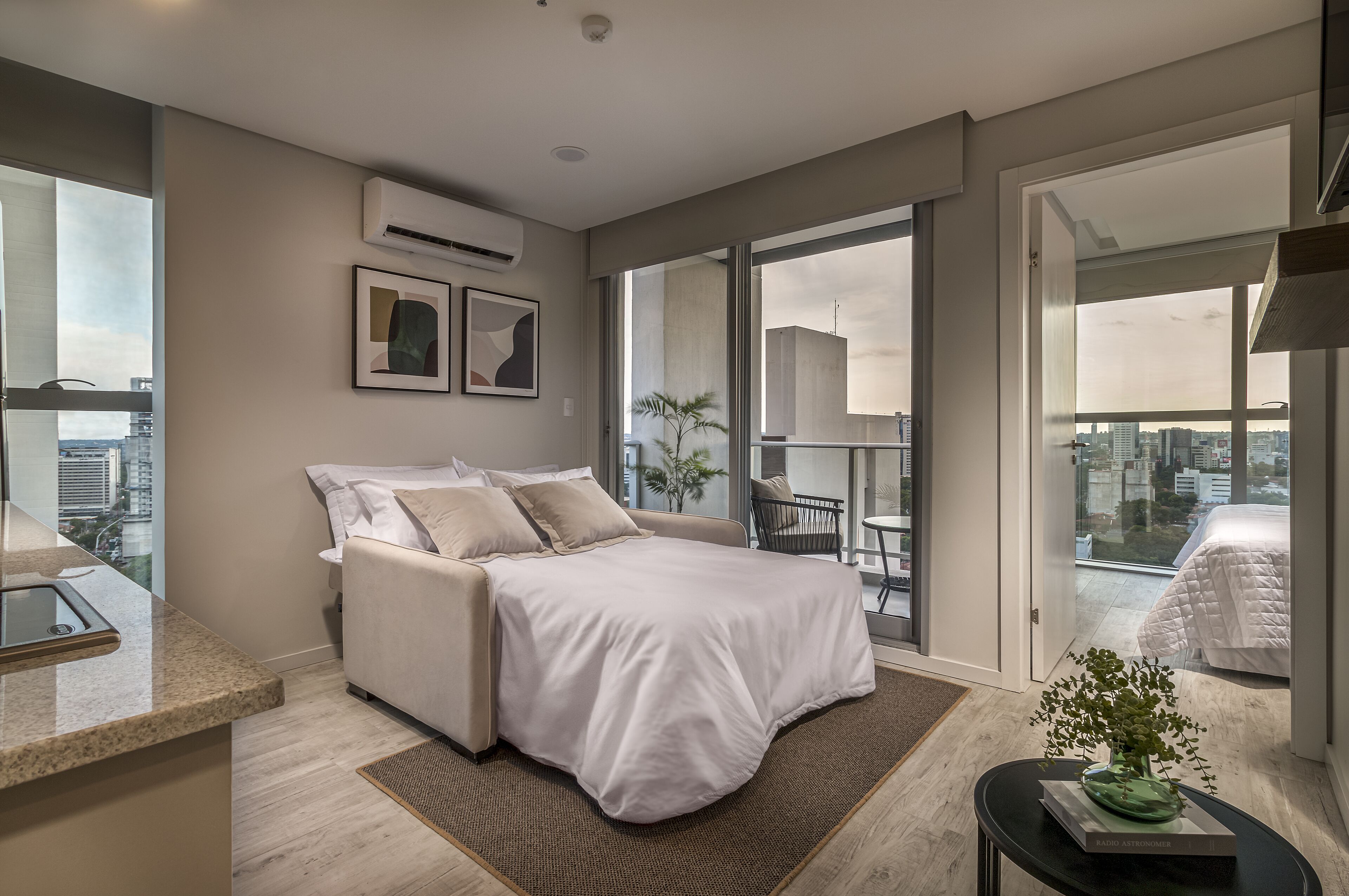 Un dormitorio moderno con una cama grande, una mesita con una planta y vistas a la ciudad a través de amplios ventanales.