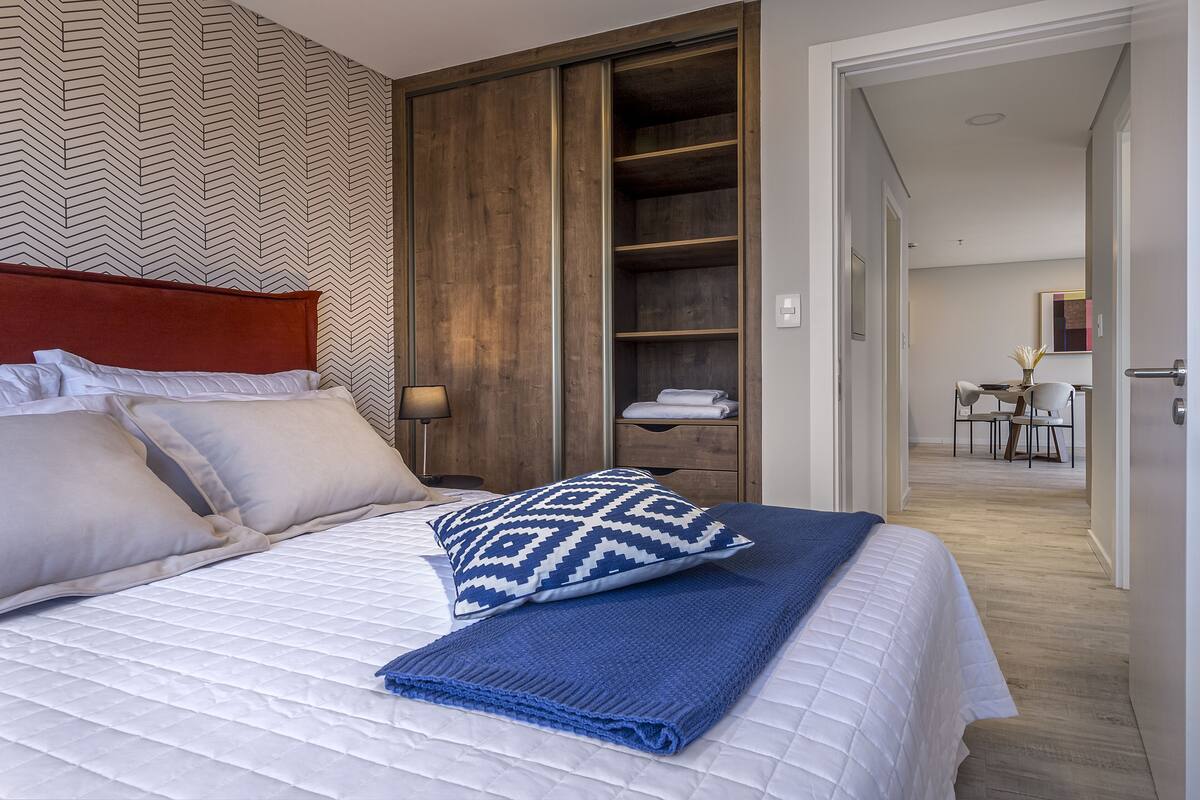 A neatly made bed with a patterned pillow, a blue blanket, and a wooden wardrobe in the background.